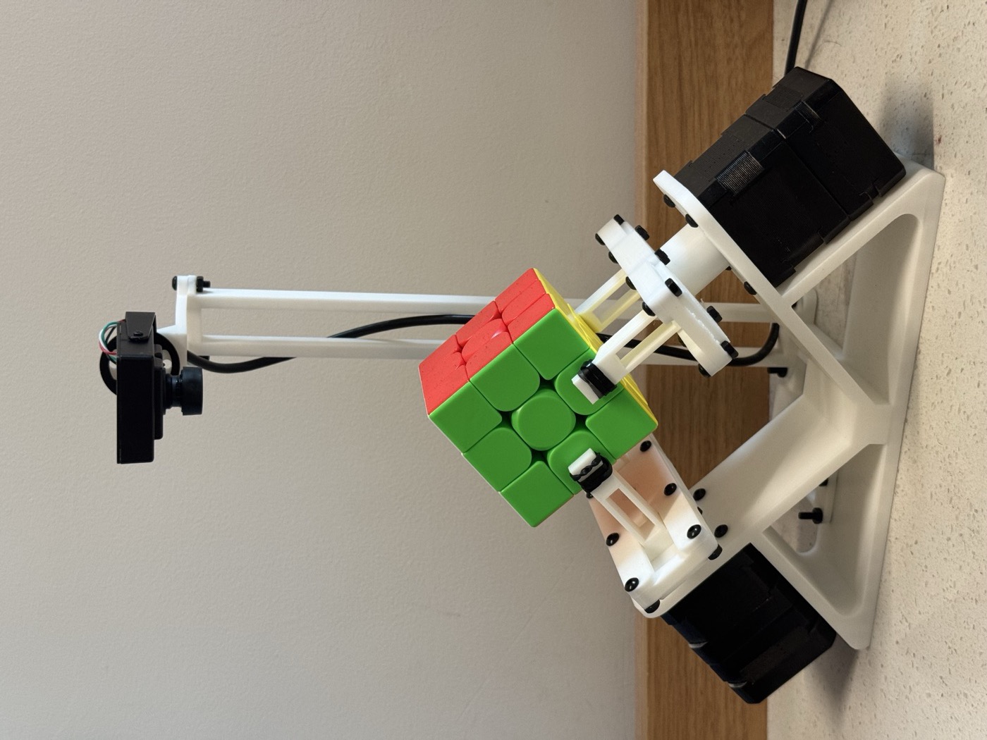 The assembled robot: white printed frame, two black stepper motors, a cube held corner-down between two grippers, and a camera on a mast above